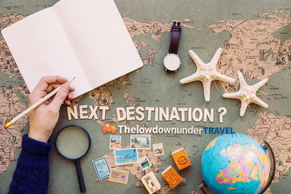 Hand holding a pencil over an open notebook on a world map, with travel items and text “Next Destination? Thelowdownunder Travel”.